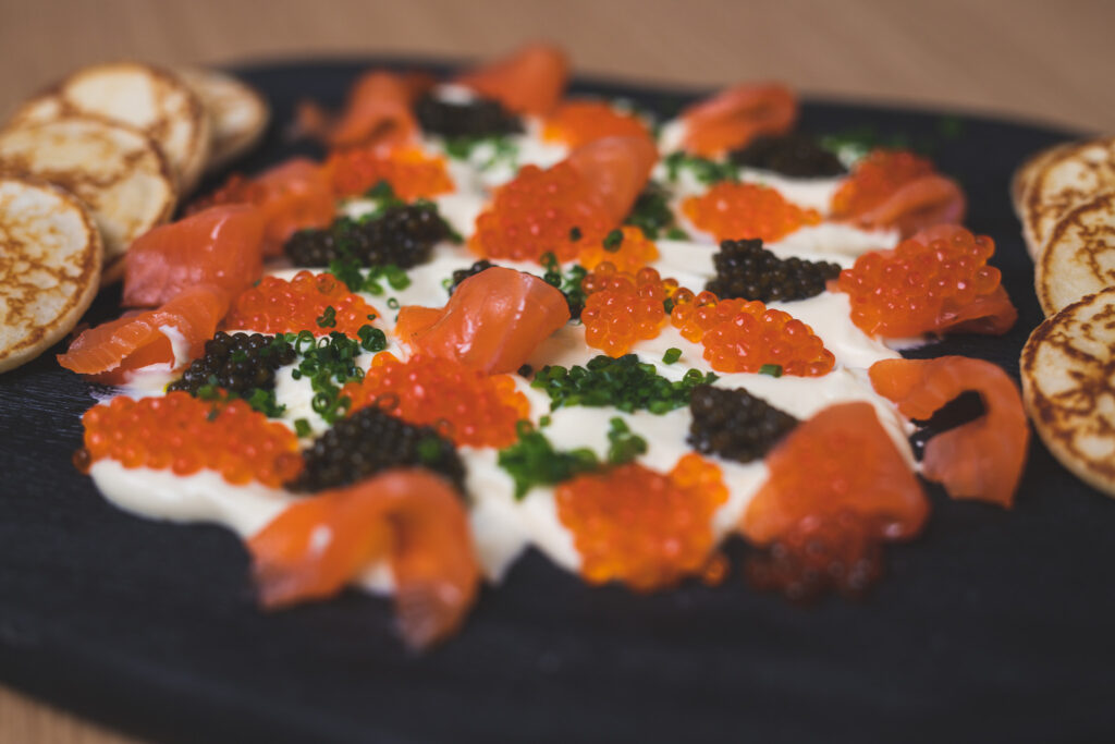 Platte mit Keta-Kaviar, schwarzem Kaviar, Räucherlachs, Schnittlauch und Crème fraîche, daneben Blinis